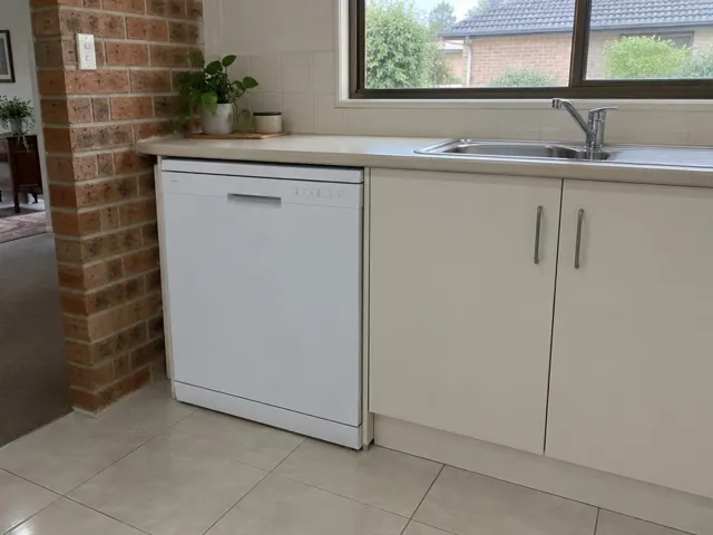 White dishwasher installed under a kitchen benchtop beside a sink, with tiled floor and brick wall in Valentine