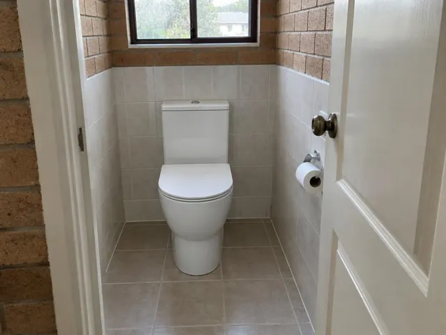 White toilet in a small tiled bathroom with brick walls and a window in Floraville